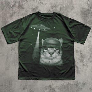 K&ouml;nnte beinhalten: Dunkelgr&uuml;nes T-Shirt mit einem schwarz-wei&szlig;en Aufdruck einer Katze mit Helm, die von einem UFO entf&uuml;hrt wird. Das Design beinhaltet Sterne. Das T-Shirt besteht aus einem weichen Material.