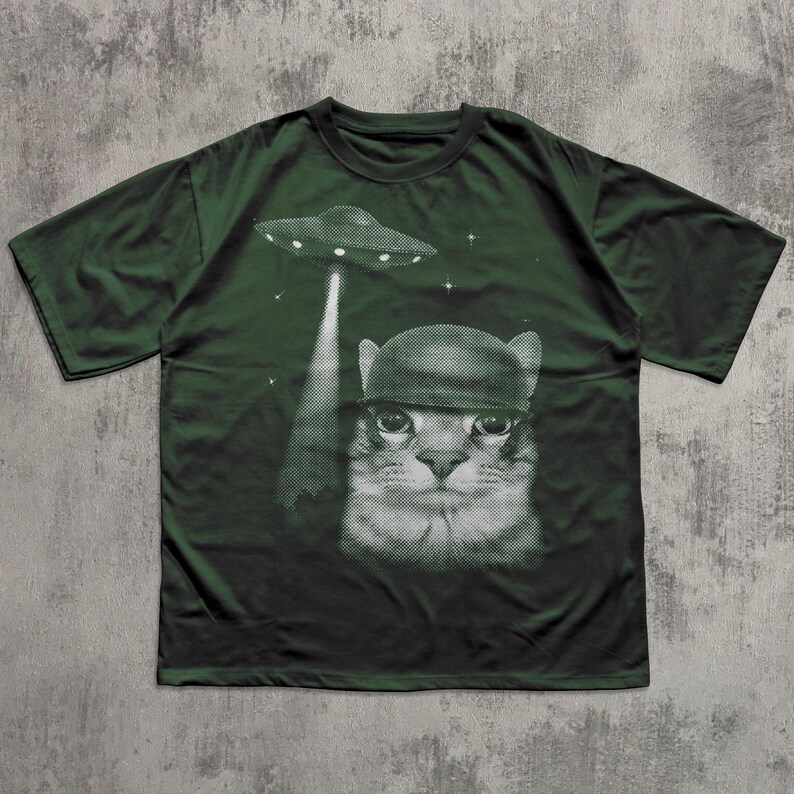 K&ouml;nnte beinhalten: Dunkelgr&uuml;nes T-Shirt mit einem schwarz-wei&szlig;en Aufdruck einer Katze mit Helm, die von einem UFO entf&uuml;hrt wird. Das Design beinhaltet Sterne. Das T-Shirt besteht aus einem weichen Material.