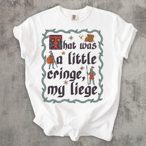 Puede incluir: Una camiseta blanca de manga corta con un estampado gr&aacute;fico que incluye el texto "That was a little cringe, my liege" en una fuente g&oacute;tica oscura. El dise&ntilde;o muestra dos caballeros, un cofre del tesoro y estrellas, dentro de un borde de enredadera.