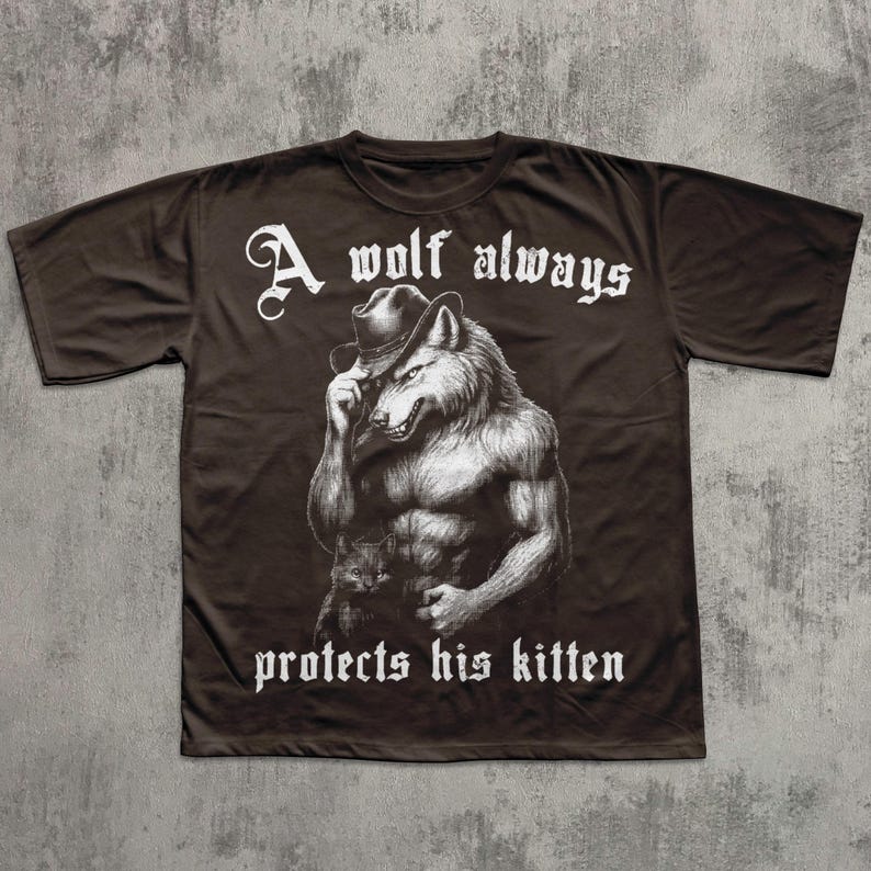 May include: Brown t-shirt featuring a detailed illustration of a muscular wolf wearing a hat, holding a kitten. The text "A wolf always protects his kitten" is printed in white, gothic-style font above and below the image.