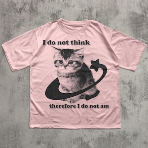 May include: A light pink t-shirt featuring a black and white graphic of a kitten encircled by a crescent and star. The text "I do not think therefore I do not am" is printed above and below the graphic in black.