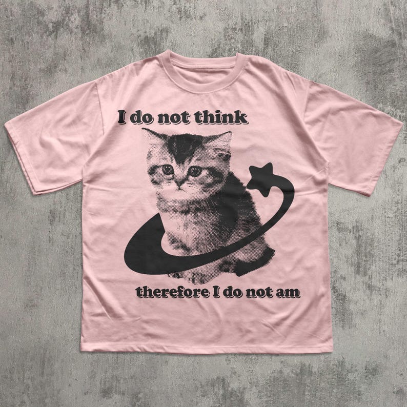 May include: A light pink t-shirt featuring a black and white graphic of a kitten encircled by a crescent and star. The text "I do not think therefore I do not am" is printed above and below the graphic in black.