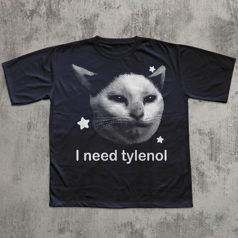 May include: Black t-shirt featuring a white, pixelated cat face with three white stars. The text "I need tylenol" is printed below the cat image. The shirt is made of a soft material.