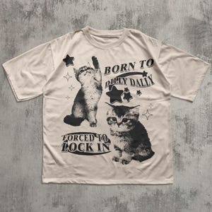 May include: A cream-coloured t-shirt with a black and white cat design and the text "BORN TO DILLY DALLY" and "FORCED TO LOCK IN". The design includes star and diamond shapes.