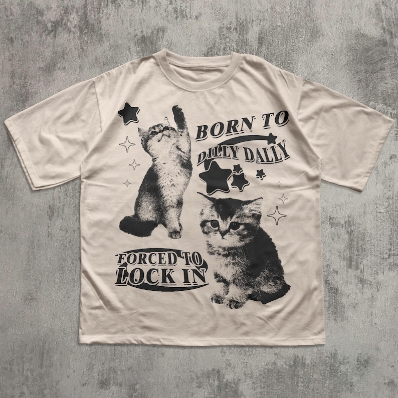 May include: A cream-coloured t-shirt with a black and white cat design and the text "BORN TO DILLY DALLY" and "FORCED TO LOCK IN". The design includes star and diamond shapes.