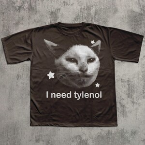 May include: Brown t-shirt featuring a white cat face graphic with stars and the text "I need tylenol". The cat graphic is a halftone design. The shirt has short sleeves and a crew neck.
