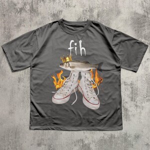 May include: Dark gray t-shirt featuring a graphic design. The design includes a fish wearing a crown above a pair of white sneakers with flames. The word "fih" is printed above the fish in white.