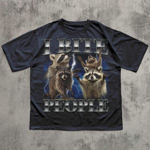 May include: Dark navy blue t-shirt featuring a graphic of four raccoons with lightning bolts and the text "I BITE PEOPLE" in a metallic silver font. The raccoons have brown and gray fur.