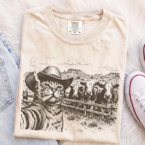 Może przedstawiać: Beżowy t-shirt z ciemnobrązowym nadrukiem graficznym przedstawiającym kota w kowbojskim kapeluszu palącego cygaro, stojącego przed drewnianym płotem z trzema krowami na tle pustynnego krajobrazu. Koszulka posiada metkę marki Comfort Colors.