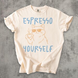 May include: Cream-colored t-shirt with the phrase "ESPRESSO YOURSELF" in blue lettering. A cat wearing sunglasses and holding a coffee cup is in the center. The shirt has short sleeves and a crew neck.