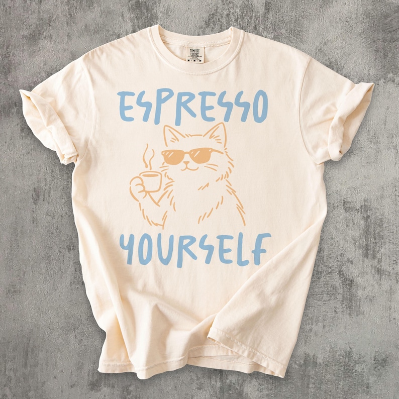 May include: Cream-colored t-shirt with the phrase "ESPRESSO YOURSELF" in blue lettering. A cat wearing sunglasses and holding a coffee cup is in the center. The shirt has short sleeves and a crew neck.