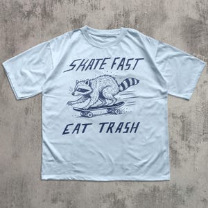 May include: Light blue t-shirt featuring a raccoon skateboarding. The raccoon is illustrated in black and white, with the words "SKATE FAST" above and "EAT TRASH" below the graphic. The shirt has short sleeves and a crew neck.