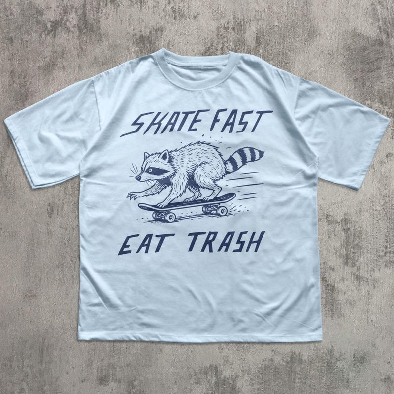 May include: Light blue t-shirt featuring a raccoon skateboarding. The raccoon is illustrated in black and white, with the words "SKATE FAST" above and "EAT TRASH" below the graphic. The shirt has short sleeves and a crew neck.
