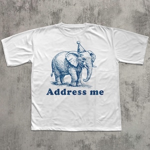 May include: White t-shirt with a blue illustration of an elephant wearing a party hat. The text "Address me" is printed below the elephant. The shirt is made of a soft material.
