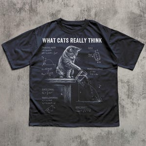 May include: A black t-shirt featuring a white graphic of a cat pushing a glass off a table, surrounded by physics equations and the text "WHAT CATS REALLY THINK". The design includes diagrams for projectile motion, kinetic energy, and impact velocity.