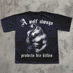 May include: Black t-shirt featuring a muscular wolf wearing a hat, holding a kitten. The text "A wolf always protects his kitten" is printed in white, gothic-style font above and below the wolf illustration.
