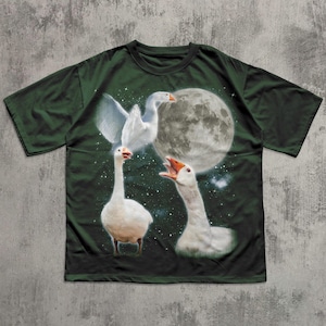Op de afbeelding: Een donkergroen t-shirt met korte mouwen en een grafische print van drie witte ganzen tegen een nachtelijke hemel. Het ontwerp bevat een grote volle maan, verspreide witte sterren, &eacute;&eacute;n gans met gespreide vleugels en twee ganzen met open snavels.