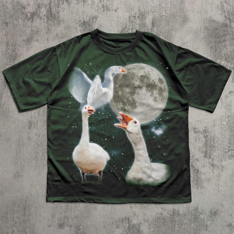 Op de afbeelding: Een donkergroen t-shirt met korte mouwen en een grafische print van drie witte ganzen tegen een nachtelijke hemel. Het ontwerp bevat een grote volle maan, verspreide witte sterren, &eacute;&eacute;n gans met gespreide vleugels en twee ganzen met open snavels.