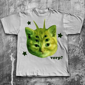 May include: A light gray t-shirt featuring a green, six-eyed cat face with antennae and green star accents. The word "vorp?" is printed in green below the cat's face. The shirt is made of a soft material.