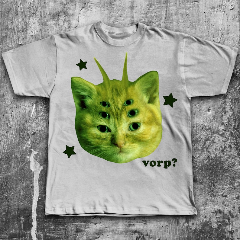May include: A light gray t-shirt featuring a green, six-eyed cat face with antennae and green star accents. The word "vorp?" is printed in green below the cat's face. The shirt is made of a soft material.