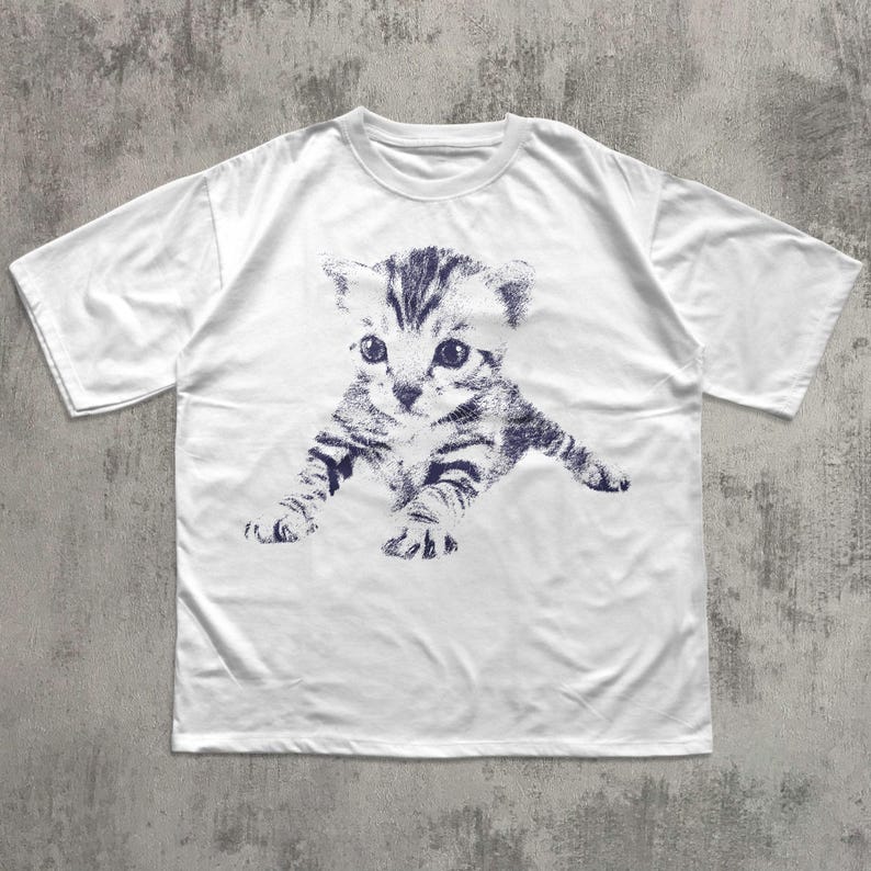 K&ouml;nnte beinhalten: Wei&szlig;es T-Shirt mit einer blauen Illustration eines K&auml;tzchens. Das K&auml;tzchen ist in einer spielerischen Pose dargestellt, mit ausgestreckten Pfoten. Die Grafik ist auf dem T-Shirt zentriert und schafft ein einfaches, aber auff&auml;lliges Design. Das T-Shirt ist aus weichem Material.