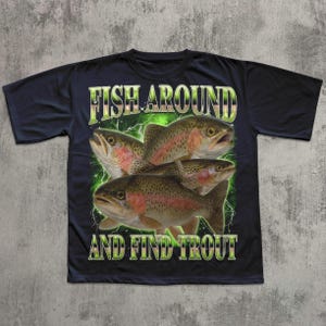 May include: Black t-shirt featuring a graphic of four trout fish with the text "FISH AROUND AND FIND TROUT" in green and white. The fish are brown with red and pink accents, set against a green lightning-like background.
