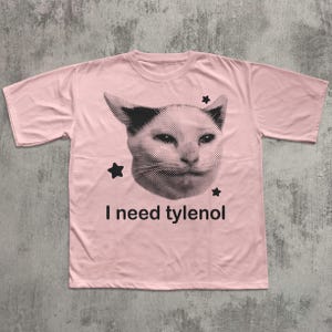 May include: A light pink t-shirt featuring a black and white image of a cat's face with three black stars. The text "I need tylenol" is printed below the cat image.