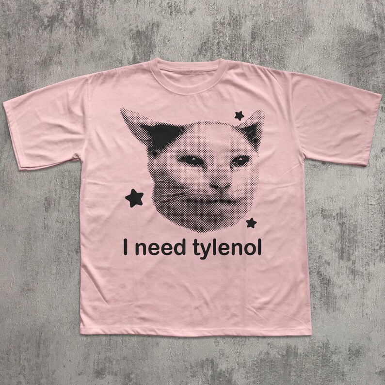 May include: A light pink t-shirt featuring a black and white image of a cat's face with three black stars. The text "I need tylenol" is printed below the cat image.