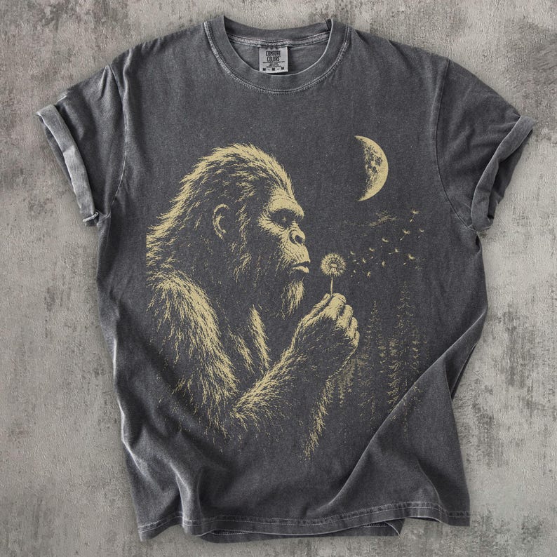 May include: A charcoal gray short-sleeve t-shirt featuring a light yellow graphic print of a hairy creature blowing on a dandelion. The design includes a crescent moon and a forest silhouette in the background. The shirt has a crew neckline.