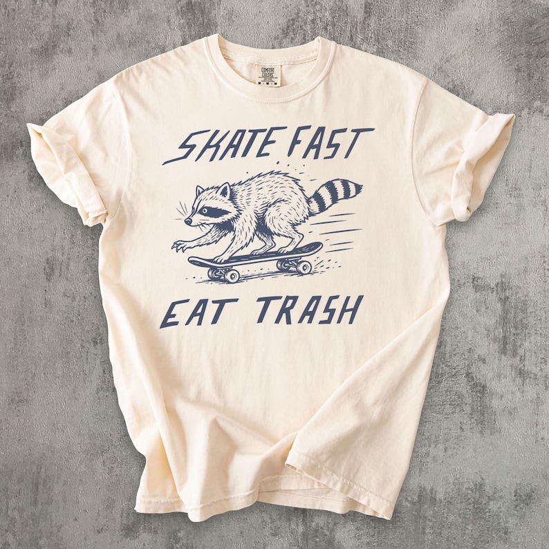 May include: Cream-colored t-shirt featuring a raccoon skateboarding. The raccoon is illustrated in navy blue, with the words "SKATE FAST EAT TRASH" above and below the graphic. The shirt has short sleeves and a crew neck.
