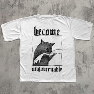 Op de afbeelding: Wit t-shirt met een zwarte afbeelding van een kat die in een hand bijt, omlijnd in een zwarte rechthoek. De woorden "become" en "ungovernable" staan boven en onder de afbeelding in gotische letters.
