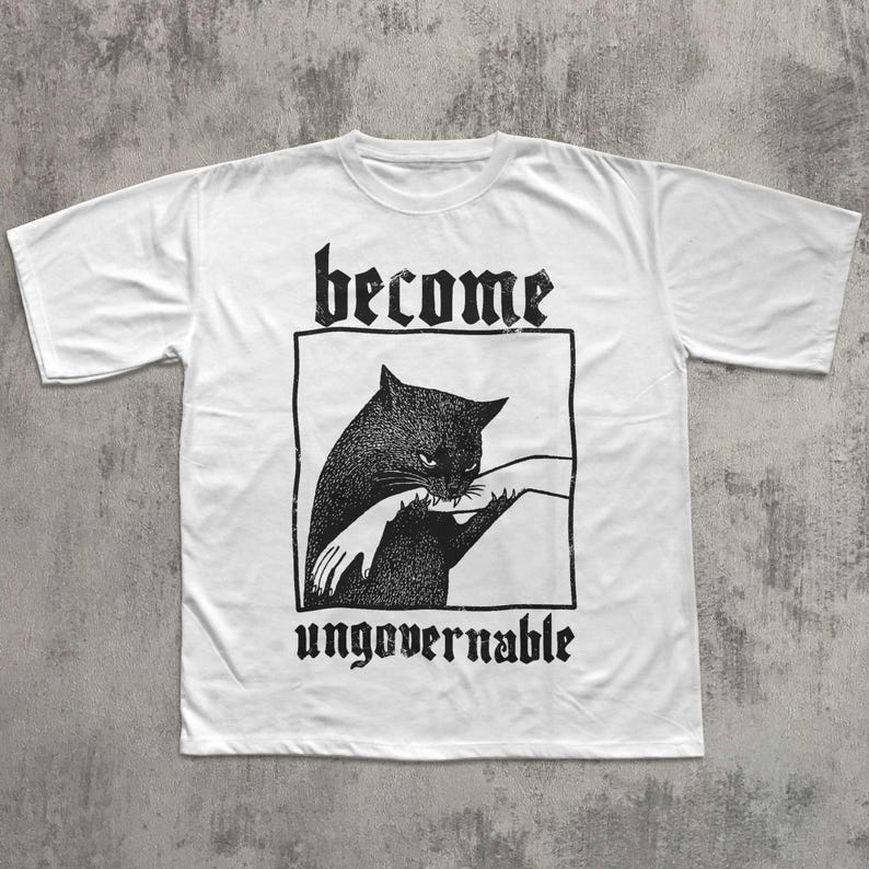 Op de afbeelding: Wit t-shirt met een zwarte afbeelding van een kat die in een hand bijt, omlijnd in een zwarte rechthoek. De woorden "become" en "ungovernable" staan boven en onder de afbeelding in gotische letters.