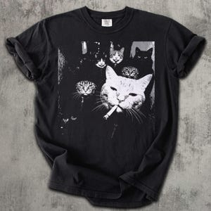May include: Black t-shirt featuring a black and white graphic of several cats. One large cat in the foreground is smoking a cigarette. The other cats are in the background.