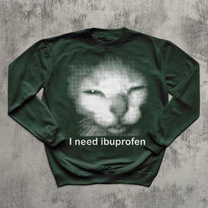 May include: Dark green sweatshirt featuring a white, halftone image of a cat's face. The text "I need ibuprofen" is printed in white below the cat image. The sweatshirt has long sleeves and a crew neck.