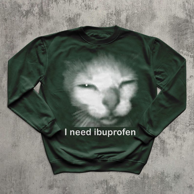 May include: Dark green sweatshirt featuring a white, halftone image of a cat's face. The text "I need ibuprofen" is printed in white below the cat image. The sweatshirt has long sleeves and a crew neck.