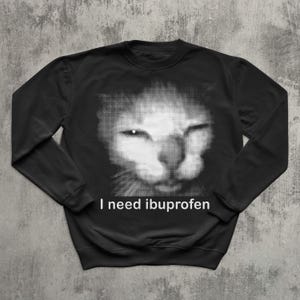 May include: Black sweatshirt featuring a grayscale image of a cat's face. The cat's face is created with a dotted pattern. The text "I need ibuprofen" is printed in white below the cat's face.