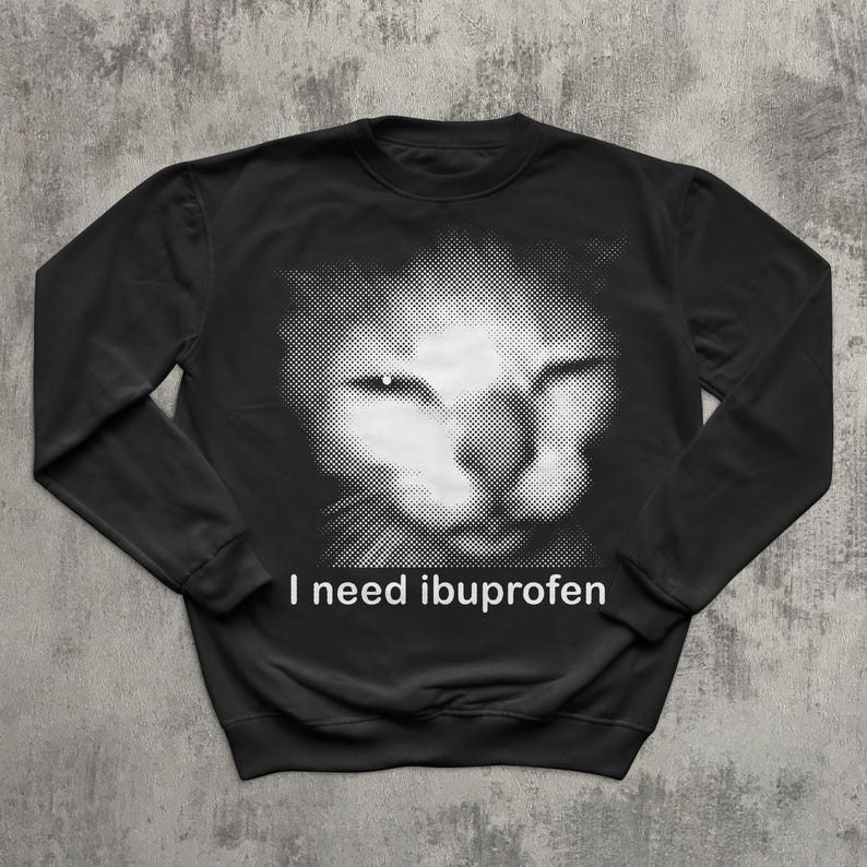 May include: Black sweatshirt featuring a grayscale image of a cat's face. The cat's face is created with a dotted pattern. The text "I need ibuprofen" is printed in white below the cat's face.