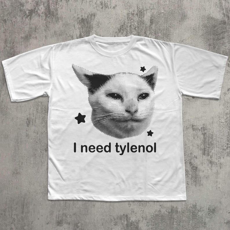 May include: White t-shirt featuring a black and white image of a cat's face with three black stars. The text "I need tylenol" is printed below the cat's face in black.