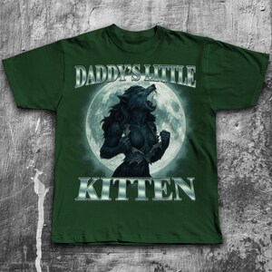 Pode incluir: T-shirt verde escuro com um gr&aacute;fico de lobisomem em frente a uma lua cheia. O texto "DADDY'S LITTLE KITTEN" &eacute; exibido acima e abaixo do gr&aacute;fico em uma fonte prateada met&aacute;lica. O lobisomem est&aacute; rasgando sua camisa.