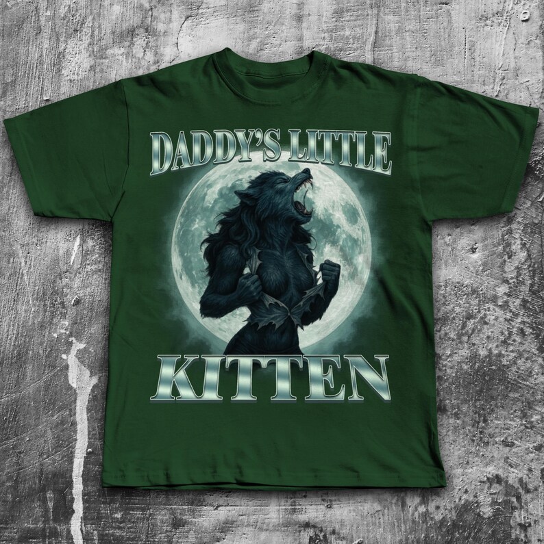 Pode incluir: T-shirt verde escuro com um gr&aacute;fico de lobisomem em frente a uma lua cheia. O texto "DADDY'S LITTLE KITTEN" &eacute; exibido acima e abaixo do gr&aacute;fico em uma fonte prateada met&aacute;lica. O lobisomem est&aacute; rasgando sua camisa.