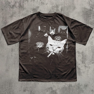 May include: Dark brown t-shirt featuring a graphic of several cat faces, with one cat smoking a cigarette. The cats are depicted in white against a dark background. The shirt has short sleeves and a crew neck.