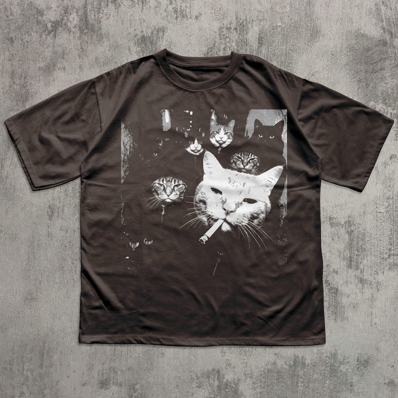 May include: Dark brown t-shirt featuring a graphic of several cat faces, with one cat smoking a cigarette. The cats are depicted in white against a dark background. The shirt has short sleeves and a crew neck.