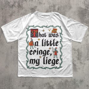 Puede incluir: Una camiseta blanca de manga corta con un estampado gr&aacute;fico y un borde decorativo de enredadera. El texto dice "That was a little cringe, my liege", acompa&ntilde;ado de ilustraciones de dos caballeros, un cofre del tesoro y estrellas.