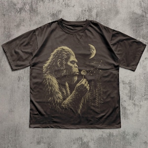 May include: A dark brown short-sleeve t-shirt featuring a graphic print of a hairy creature resembling a Bigfoot holding a dandelion. The design includes a crescent moon and a forest silhouette in the background, rendered in a light tan color.