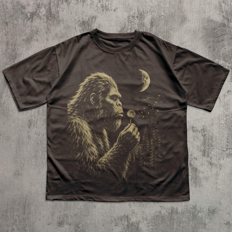 May include: A dark brown short-sleeve t-shirt featuring a graphic print of a hairy creature resembling a Bigfoot holding a dandelion. The design includes a crescent moon and a forest silhouette in the background, rendered in a light tan color.