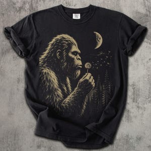 May include: A black short-sleeve t-shirt featuring a graphic print of a hairy creature in profile holding a dandelion. The design includes a crescent moon, a starry night sky, and a forest of pine trees in a muted tan color against the black fabric.