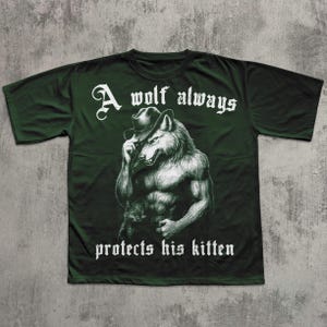 May include: Dark green t-shirt featuring a muscular wolf wearing a hat, holding a kitten. The text "A wolf always protects his kitten" is printed in white, gothic-style font. The shirt has short sleeves and a crew neck.