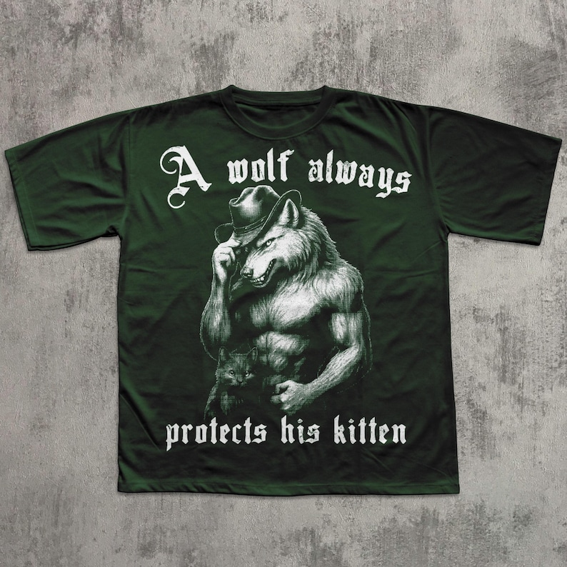 May include: Dark green t-shirt featuring a muscular wolf wearing a hat, holding a kitten. The text "A wolf always protects his kitten" is printed in white, gothic-style font. The shirt has short sleeves and a crew neck.