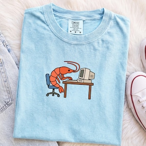 May include: A light blue t-shirt featuring a graphic of an orange shrimp sitting in a blue office chair at a brown desk, typing on a vintage computer. The shirt has a white neck label with the text Comfort Colors printed in black.