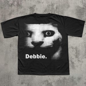 May include: Black t-shirt with a grayscale image of a face with large eyes and the name "Debbie" in white text below. The graphic is a pixelated design, creating a unique visual effect.